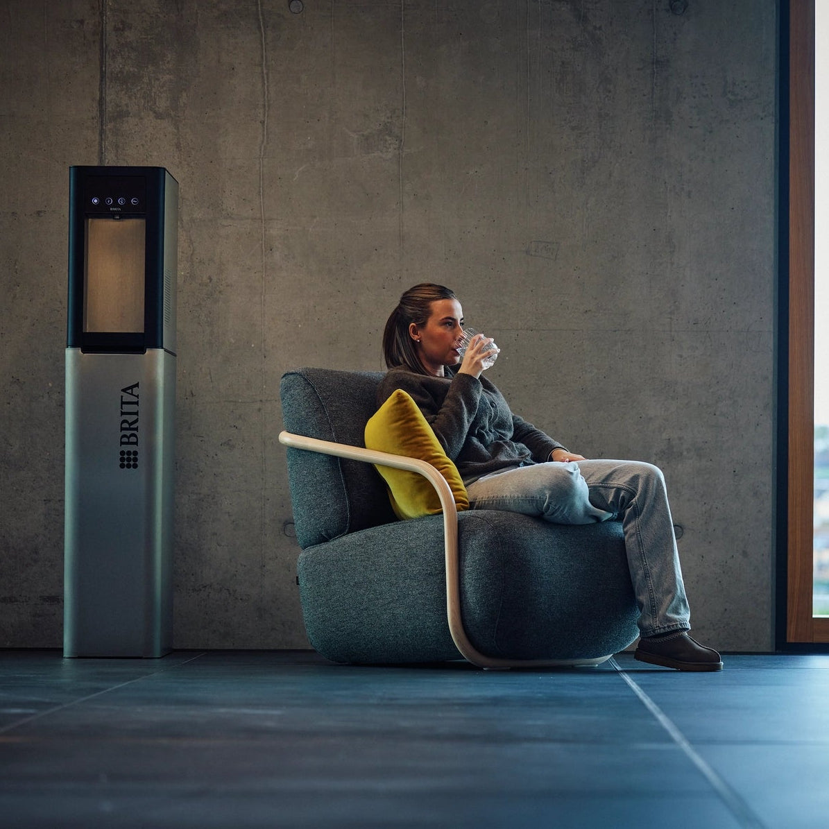 A woman sits in a modern gray armchair with a yellow pillow, drinking from a cup, next to a tall silver and black BRITA Top Pro water dispenser in a minimalist room with a large window overlooking a city.