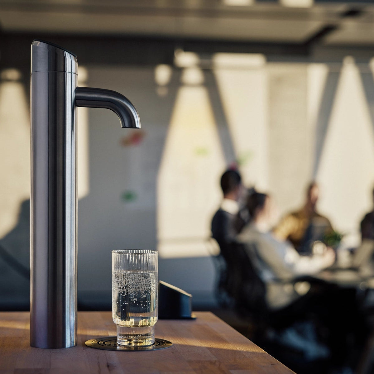 Een BRITA Extra I-Tap-waterdispenser op een houten tafel, met daaronder een glas water, in een wazige kantooromgeving.