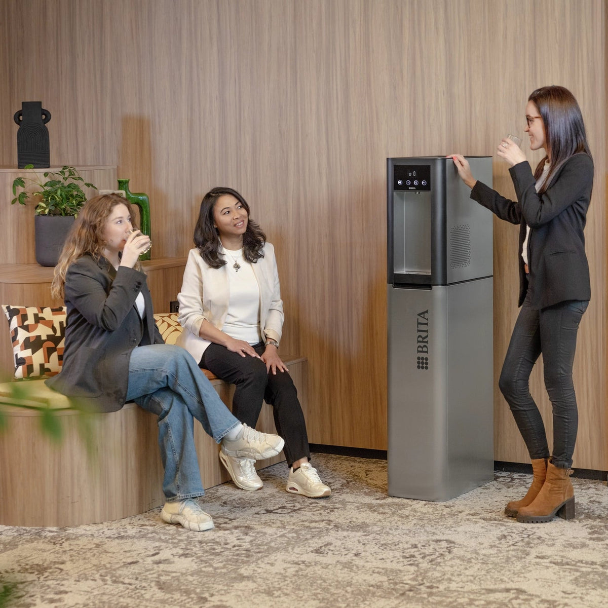 Een vrouw die naast een Top Pro-waterdispenser staat in een moderne kantooromgeving, terwijl twee andere vrouwen op een bankje zitten.