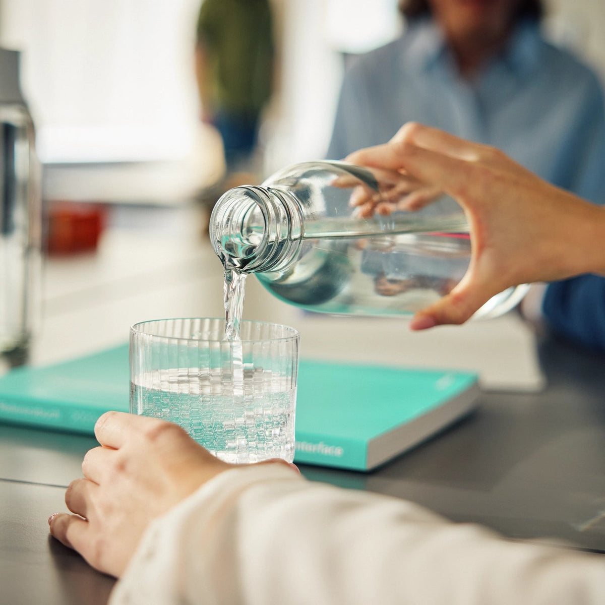 Iemand die water uit een fles in een glas giet dat op een kantoortafel staat.